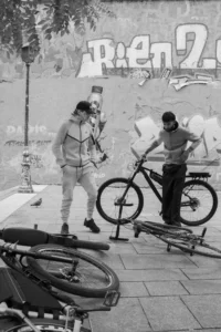 Cette photographie en noir et blanc montre deux hommes près de vélos. Le sujet principal semble être l’un d’eux, en train d’observer ou de discuter avec l’autre, qui est penché sur un vélo.La composition est simple et naturelle, avec un cadrage serré sur les personnages et les vélos. La lumière est douce, sans ombres marquées, créant une ambiance calme et quotidienne. Les couleurs, bien que monochromes, jouent sur les contrastes de tons clairs et foncés.Le style est documentaire, évoquant une scène de rue authentique et spontanée. L’ambiance est sobre et réaliste, renforcée par le flou des éléments en arrière-plan.