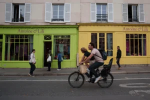 Cette photographie met en scène un couple à vélo comme sujet principal, au premier plan. La composition joue sur la profondeur avec les passants en arrière-plan devant deux bâtiments colorés, l’un vert et l’autre jaune, portant l’enseigne "Antoine & Lili".Le cadrage est serré sur les cyclistes, avec une perspective dynamique. La lumière est naturelle, légèrement tamisée, créant des ombres douces. Les couleurs dominantes sont vives et contrastées : vert, jaune, gris et noir.Le style est documentaire et urbain, capturant un moment de vie quotidienne. L’ambiance est vivante et animée, reflétant une scène typique d’une ville européenne.