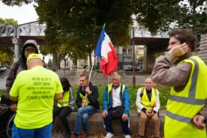 Cette photographie montre un groupe de manifestants assis sur des barricades. Le sujet principal est un homme tenant un drapeau français, entouré de personnes vêtues de gilets jaunes, symboles des Gilets Jaunes.La composition est dynamique, avec un cadrage serré sur les manifestants. La lumière naturelle éclaire la scène, mettant en valeur les visages et les couleurs vives. Les couleurs dominantes sont le jaune des gilets, le bleu-blanc-rouge du drapeau, et les tons neutres des vêtements.Le style est documentaire, capturant un moment authentique. L’ambiance est à la fois solennelle et combative, reflétant un esprit de protestation.
