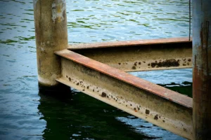 Cette photographie met en scène un **ponton en béton et métal rouillé** partiellement immergé dans l’eau. Le **sujet principal** est ce support érodé, soulignant son usure et son contact avec l’eau.La **composition** est épurée, avec un **cadrage serré** centré sur les éléments structurels. La **lumière**, douce et naturelle, crée des contrastes entre ombres et reflets. Les **couleurs dominantes** sont les tons gris-beige du béton, le rouge rouille du métal et les bleus-verts de l’eau.Le **style** est réaliste et documentaire, évoquant une **ambiance mélancolique et industrielle**, presque abandonnée.