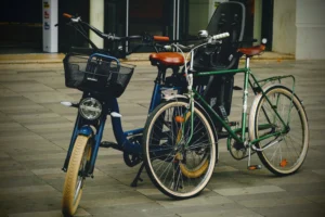 Cette photographie met en scène deux vélos stationnés côte à côte. Le sujet principal est un vélo électrique bleu avec panier avant, accompagné d’un vélo classique vert à l’arrière.La composition est symétrique et épurée, avec un cadrage serré sur les vélos. La lumière, tamisée et légèrement artificielle, crée des ombres douces, évoquant une ambiance nocturne ou crépusculaire. Les couleurs dominantes sont les teintes bleutées, vertes et grises, renforçant une atmosphère calme et moderne. Le style est sobre et urbain, tandis que l’ambiance, à la fois fonctionnelle et poétique, suggère une pause dans la ville.
