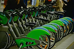 Cette photographie montre une rangée de vélos en libre-service, probablement des vélos de type "Vélib’". Le sujet principal est ces vélos alignés, avec des couleurs dominantes vertes et bleues. La composition est linéaire et ordonnée, avec un cadrage serré sur les vélos, mettant en valeur leur design moderne.La lumière semble naturelle et douce, créant des ombres légères. L’ambiance est urbaine et pratique, évoquant un système de transport partagé. Le style est réaliste et documentaire, captant une scène du quotidien.