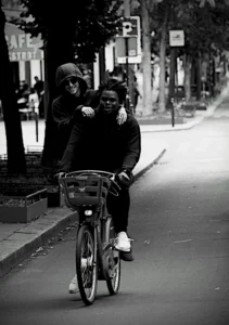 Cette photographie en noir et blanc met en scène un couple à vélo comme sujet principal. Le cadrage est serré, centré sur les deux personnes, avec un léger flou d’arrière-plan.La composition est dynamique, avec une ligne de fuite créée par la route et les arbres alignés. La lumière semble naturelle, douce, et crée des contrastes modérés. L’ambiance est intime et nostalgique, renforcée par le style rétro du cliché. Les couleurs dominantes, bien que monochromes, jouent sur les nuances de gris et de noir, accentuant l’émotion et la simplicité du moment.