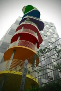 Cette photographie met en scène une structure architecturale colorée et moderne, probablement un escalier extérieur ou une tour d'accès. Le sujet principal est cette tour aux couleurs vives (orange, jaune, rouge, vert, bleu), disposées en cercles superposés.La composition est verticale, avec un cadrage serré qui accentue la hauteur de la structure. La lumière est naturelle, légèrement diffuse, créant des contrastes doux. Les couleurs dominantes sont vives et saturées, apportant une touche joyeuse et dynamique. Le style est contemporain et artistique, évoquant une ambiance ludique et innovante dans un environnement urbain.