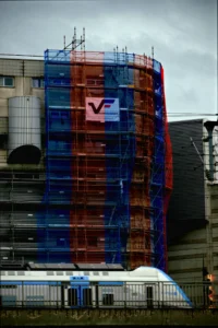 Cette photographie montre un immeuble en cours de rénovation, recouvert de filets de protection colorés (bleu, rouge et transparent). Le sujet principal est le bâtiment avec un logo "V" visible. La composition met en avant la verticalité et la structure architecturale.Le cadrage est serré, centré sur l'immeuble. La lumière est diffuse, probablement par temps nuageux, créant une ambiance sobre. Les couleurs dominantes sont les teintes bleues et rouges des filets, contrastant avec le gris du ciel et des bâtiments environnants. Le style est documentaire et réaliste, évoquant une ambiance urbaine et industrielle.
