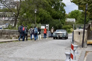 Cette photographie montre un groupe de personnes marchant sur un chemin en pente près d’un véhicule ancien. Le sujet principal est ce groupe, composé de plusieurs individus habillés de manière décontractée.La composition est dynamique, avec un cadrage légèrement bas, mettant en valeur les personnes et le véhicule. La lumière est naturelle, légèrement diffuse, créant des ombres douces. Les couleurs dominantes sont les tons neutres (gris, beige) avec des touches de vert (végétation) et bleu (ciel).Le style est documentaire, capturant une scène du quotidien. L’ambiance est calme et un peu nostalgique, renforcée par le véhicule rétro et l’environnement urbain paisible.