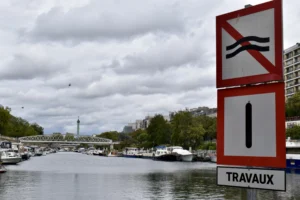 Cette photographie montre un panneau de signalisation indiquant des travaux ("TRAVAUX") avec des symboles de restrictions (accès interdit aux piétons et véhicules).La composition met en avant le panneau en premier plan, tandis que l’arrière-plan révèle un canal avec des bateaux et des bâtiments. Le cadrage est serré sur le panneau, mais inclut une vue dégagée sur le paysage urbain.La lumière est douce, légèrement voilée par des nuages. Les couleurs dominantes sont les tons gris-bleu du ciel et de l’eau, contrastant avec le rouge vif du panneau. L’ambiance est calme et urbaine, évoquant une journée ordinaire en ville.