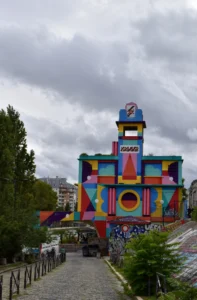 Cette photographie met en scène un bâtiment coloré et mural, couvert de fresques géométriques et psychédéliques. Le sujet principal est cette structure artistique, aux formes cubiques et aux couleurs vives.La composition inclut une rue en pente avec des escaliers et des passants. Le cadrage est serré sur le bâtiment, encadré par des arbres et des éléments urbains.La lumière est naturelle, un peu grise à cause du ciel couvert. Les couleurs dominantes sont vives et contrastées : bleus, rouges, jaunes et verts.Le style est artistique et street art, créant une ambiance joyeuse et dynamique malgré le temps nuageux.