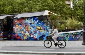 Cette photographie met en scène un bus décoré de fresques colorées représentant des personnages de films et séries cultes. Le sujet principal est un cycliste passant devant ce bus.La composition est dynamique, avec un cadrage serré sur le vélo et le bus. La lumière est naturelle et semble provenir d’un jour ensoleillé. Les couleurs dominantes sont vives et variées, allant du bleu au rouge, en passant par des teintes pastel, créant un style pop-art et festif. L’ambiance est joyeuse et énergique, évoquant une célébration de la culture populaire.