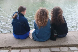 Cette photographie montre trois jeunes femmes assises sur une margelle de pierre, le dos tourné à la caméra, face à l’eau. Le **sujet principal** est leur posture détendue et leur interaction silencieuse.La **composition** est simple et symétrique, avec un **cadrage serré** centré sur elles. La **lumière** semble naturelle, douce, sans ombres marquées, évoquant un jour nuageux. Les **couleurs dominantes** sont les tons bleus (vêtements) et gris (pierres, eau), adoucis par des nuances naturelles.Le **style** est réaliste et documentaire, capturant un moment intime. L’**ambiance** est calme et contemplative, presque mélancolique.