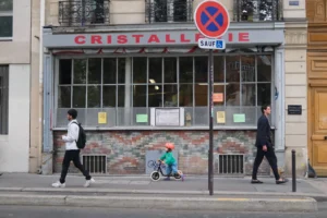 Cette photographie montre une rue urbaine devant un magasin nommé **« Cristallerie »**. Le sujet principal est une scène de vie quotidienne : trois personnes (dont un enfant sur un trottinette) traversent ou marchent près d’un passage piéton.La composition est dynamique, avec un cadrage serré sur le magasin et ses vitrines. La lumière est naturelle, légèrement tamisée, créant des ombres douces. Les couleurs dominantes sont les tons gris (bâtiments), verts (enseignes) et bleus (ciel). L’ambiance est calme et réaliste, reflétant une ville animée mais paisible. Le style est documentaire et spontané.