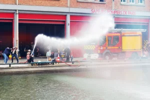 Cette photographie montre un **camion de pompiers** en action, projetant un puissant jet d’eau. Le **sujet principal** est l’intervention des secours, avec des pompiers et spectateurs en arrière-plan.La **composition** est dynamique, centrée sur le jet d’eau et le véhicule. Le **cadrage** est serré, mettant en valeur l’action. La **lumière** semble naturelle, bien que légèrement contrastée.Les **couleurs dominantes** sont les tons rouges (bâtiment), jaunes (véhicule) et gris (eau). L’**ambiance** est tendue et active, reflétant une situation d’urgence. Le style est documentaire, réaliste et immédiat.
