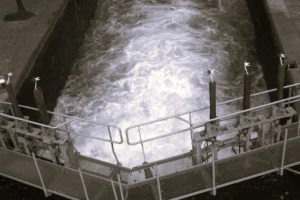 Cette photographie montre une **piscine d’eau agitée**, probablement un bassin industriel ou une usine de traitement.Le **sujet principal** est l’eau en mouvement turbulent, avec des remous visibles. La **composition** est centrée, avec un cadrage serré sur le bassin entouré de **barrières métalliques**. La **lumière** est artificielle, froide et uniforme, créant des contrastes nets. Les **couleurs dominantes** sont des teintes grises et blanches, renforçant une ambiance industrielle et technique. Le style est **documentaire et fonctionnel**, évoquant une ambiance **austère et mécanique**.