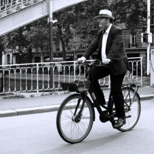Cette photographie en noir et blanc met en scène un homme en costume élégant, chapeau et cravate, comme sujet principal. Il est assis sur un vélo classique, en mouvement.La composition est simple et équilibrée, avec une perspective légèrement basse. Le cadrage isole le sujet sur la route, encadré par une rambarde et des arbres en arrière-plan.La lumière semble naturelle, douce, créant des contrastes modérés. Les couleurs dominantes sont les nuances de gris typiques du noir et blanc.Le style évoque une ambiance rétro, intemporelle et sophistiquée, rappelant les époques passées. L'ambiance est calme et élégante.