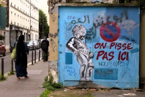 Cette photographie montre un mur tagué avec des graffitis en français et en arabe, dont la phrase *« On pisse pas ici »*. Le sujet principal est le graffiti, avec une silhouette stylisée et des symboles politiques.La composition inclut deux personnes en arrière-plan, floutées, marchant sur un trottoir urbain. Le cadrage est serré sur le mur, avec une perspective légèrement basse. La lumière est naturelle, diffuse, typique d’un jour couvert. Les couleurs dominantes sont les tons bleus, blancs et gris, renforçant une ambiance urbaine et légèrement mélancolique, mêlant protestation et vie quotidienne.