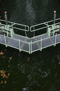 Cette photographie montre un ponton métallique flottant sur l’eau, sujet principal. La composition est symétrique avec des barrières latérales en fil métallique. Le cadrage est serré, mettant en valeur la structure et son reflet sur l’eau.La lumière semble naturelle, probablement prise en journée avec un éclairage doux. Les couleurs dominantes sont les tons gris métallique du ponton, contrastant avec le noir de l’eau et les feuilles mortes en arrière-plan.Le style est réaliste et documentaire, créant une ambiance calme et légèrement mélancolique.