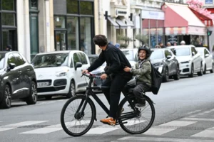Cette photographie met en scène deux personnes à vélo en ville. Le sujet principal est le cycliste en premier plan, vêtu d’un manteau sombre, avec un passager assis derrière lui.La composition montre une rue urbaine avec des voitures en arrière-plan et des bâtiments. Le cadrage est serré sur les cyclistes, capturant leur mouvement.La lumière semble naturelle, probablement en journée, avec des ombres douces. Les couleurs dominantes sont les tons gris et neutres, adoucis par des touches de vert et de rouge (voiture et enseigne).Le style est documentaire et réaliste, évoquant une ambiance dynamique et quotidienne en milieu urbain.