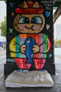 Cette photographie montre un personnage stylisé peint sur un support vertical, ressemblant à un mélange de bonhomme et de poisson. Le sujet principal est un dessin coloré et naïf, avec des traits expressifs et des couleurs vives.La composition est centrée sur ce personnage, encadré par une bordure colorée. Le cadrage est serré, mettant en valeur les détails. La lumière semble naturelle, éclairant uniformément la scène. Les couleurs dominantes sont vives et contrastées : rouge, bleu, jaune et vert.Le style évoque l’art urbain ou street art, avec une touche enfantine et joyeuse, créant une ambiance ludique et festive.