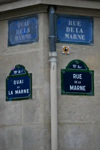 Cette photographie montre deux plaques de rue parisiennes indiquant "Quai de la Marne" et "Rue de la Marne". Le sujet principal est ces plaques bleues et blanches, fixées sur un mur en pierre grise.La composition est symétrique, avec un cadrage serré mettant en valeur les détails. La lumière naturelle éclaire uniformément la scène, sans ombre marquée. Les couleurs dominantes sont le bleu des plaques, le gris de la pierre et le noir des éléments métalliques. Le style est documentaire et urbain, évoquant une ambiance typiquement parisienne, sobre et classique.