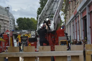 Cette photographie montre un pompier en action, utilisant une échelle de secours pour atteindre un bâtiment. Le sujet principal est ce professionnel, vêtu d'une tenue noire, en train de monter l'échelle.La composition met en avant l'échelle centrale, encadrée par des véhicules de secours rouges et des spectateurs en arrière-plan. Le cadrage est serré, mettant l'accent sur l'action et l'urgence. La lumière est naturelle, légèrement voilée par des nuages, créant une ambiance réaliste et dynamique.Les couleurs dominantes sont les tons rouges des véhicules et les gris/beiges des bâtiments, avec des touches de vert des arbres. Le style est documentaire, reflétant une scène authentique et intense.