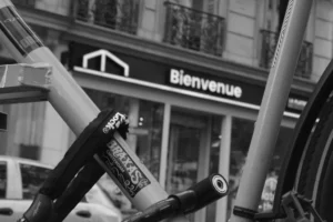 Cette photographie en noir et blanc met en avant un vélo en premier plan, partiellement cadré, avec ses roues et cadre bien visibles. L’arrière-plan montre une façade urbaine avec l’enseigne "Bienvenue" d’un magasin. La composition joue sur le contraste entre le flou de l’arrière-plan et le vélo net, créant une profondeur.La lumière semble naturelle, probablement en journée, avec des ombres douces. Les tons dominants sont les gris et noirs, typiques du noir et blanc, renforçant une ambiance urbaine et intemporelle. Le style est documentaire et minimaliste, évoquant une scène de vie quotidienne.