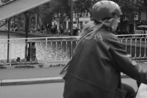 Cette photographie en noir et blanc met en scène un cycliste en premier plan, vêtu d’une veste longue et d’un casque, dont le visage est flou. La composition joue sur la profondeur avec une barrière métallique et un espace urbain en arrière-plan.Le cadrage serré isole le sujet tout en intégrant des éléments architecturaux et des passants. La lumière, douce et diffuse, crée des contrastes modérés, accentuant les textures. L’ambiance est à la fois dynamique et mélancolique, renforcée par le monochrome. Le style rappelle un documentaire urbain intemporel, évoquant une atmosphère calme et quotidienne.