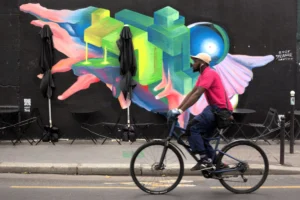 Cette photographie met en scène un cycliste comme sujet principal, vêtu d’un t-shirt rouge et d’un masque, en mouvement devant un mur. La composition joue sur le contraste entre le sujet dynamique et le fond statique.Le cadrage est serré sur le cycliste, avec une partie du mur coloré en arrière-plan. La lumière semble naturelle, bien répartie, sans ombre marquée. Les couleurs dominantes sont vives : bleu, vert, rouge et jaune sur le mural, créant un style urbain et artistique. L’ambiance est à la fois moderne et énergique, mêlant mouvement et créativité.