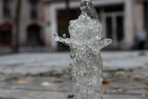 Cette photographie met en scène un **jet d’eau** en mouvement, sujet principal, formant une silhouette abstraite évoquant une figure humaine. Le **cadrage serré** isole ce détail, créant un effet dynamique. L’arrière-plan flou, avec des bâtiments et des fenêtres, suggère un espace urbain.La **lumière** semble naturelle, avec des reflets sur l’eau, et les **couleurs dominantes** sont des teintes froides (bleutées, grises). Le style est **minimaliste et épuré**, privilégiant la forme et le mouvement. L’ambiance est **poétique et contemplative**, mêlant abstraction et réalité.