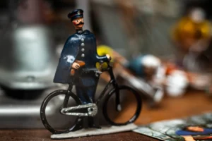 Cette photographie met en scène une figurine de policier miniature en uniforme du XIXe siècle, assis sur un vélo. Le sujet principal est net et centré, avec un cadrage serré qui isole la figurine.L'arrière-plan est flou, créant un effet de profondeur. La lumière douce et naturelle accentue les détails de la figurine. Les couleurs dominantes sont les tons sombres (bleu, noir) de l'uniforme, contrastant avec le métal argenté du vélo.Le style évoque une ambiance nostalgique et miniaturiste, renforcée par l'aspect vintage et le flou artistique de l'arrière-plan.