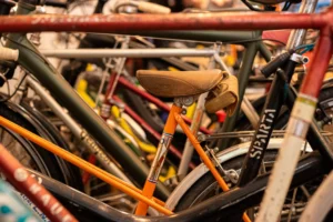 Cette photographie montre un **ensemble de vélos** entreposés, formant le sujet principal. La **composition** est serrée et désordonnée, avec des cadres et roues entrelacés. Le **cadrage** est rapproché, mettant en valeur les détails techniques et les couleurs variées.Les **couleurs dominantes** sont des tons métalliques (noir, gris, argent) et des touches de rouge, orange et vert. La **lumière** semble artificielle, légèrement tamisée, créant des contrastes doux. Le style est **documentaire et réaliste**, évoquant une ambiance **urbaine et industrielle**, presque intimiste par son désordre organisé.