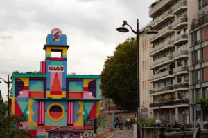 Cette photographie met en scène un bâtiment coloré et mural, couvert de fresques géométriques et vives. Le sujet principal est cette structure artistique, contrastant avec les immeubles gris en arrière-plan.La composition est dynamique, avec un cadrage serré sur la façade colorée, encadrée par des arbres et des bâtiments. La lumière est naturelle, légèrement tamisée, créant des ombres douces.Les couleurs dominantes sont vives : rose, bleu, jaune et vert, dans un style géométrique et joyeux. L’ambiance est festive et artistique, évoquant un mélange d’urbanité et de créativité.