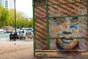 Cette photographie met en scène un **mur tagué** comme sujet principal, avec un graffiti stylisé représentant un visage. La composition joue sur le contraste entre le graffiti coloré et le cadre vert métallique. Le cadrage est serré sur le mur, tout en laissant voir un espace urbain en arrière-plan.La lumière semble naturelle, légèrement tamisée, créant des ombres douces. Les **couleurs dominantes** sont les tons vifs du graffiti (bleu, rouge, orange) et les teintes terreuses des briques. Le style est urbain et street art, évoquant une ambiance **vivante et rebelle**, mêlant créativité et négligence.