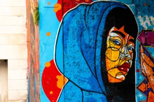 Cette photographie met en scène un **mural coloré** représentant un visage féminin stylisé, coiffé d'une capuche bleue. Le **sujet principal** est ce portrait expressif, aux traits marqués et aux couleurs vives.La **composition** est serrée, avec un **cadrage rapproché** qui accentue les détails. Les **couleurs dominantes** sont le bleu, le rouge et le jaune, créant un contraste dynamique. Le **style** est graphique et artistique, évoquant l’art urbain.L’**ambiance** est à la fois mystérieuse et énergique, renforcée par les jeux de lumière et les ombres sur les contours. L’éclairage naturel met en valeur les teintes intenses.