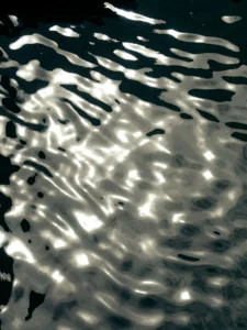 Cette photographie capture des **reflets déformés** sur une surface liquide, probablement de l’eau. Le **sujet principal** semble être les motifs abstraits créés par la lumière traversant les nuages ou un objet translucide.La **composition** est épurée, avec un **cadrage serré** qui met en valeur les jeux de lumière. La **lumière**, douce et naturelle, crée des contrastes subtils entre clarté et ombres. Les **couleurs dominantes** sont des teintes grises et blanches, évoquant une ambiance calme et presque onirique.Le **style** est minimaliste et abstrait, rappelant l’art photographique expérimental. L’**ambiance** est apaisante, presque hypnotique, comme une vue aérienne ou sous-marine.