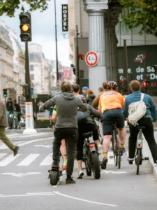 Cette photographie capture un groupe de cyclistes et de trottinetteurs traversant une intersection en ville. Le **sujet principal** est ce flux de personnes en mouvement, vu de dos.La **composition** est dynamique, avec un **cadrage serré** sur les individus, créant un effet de profondeur. La **lumière** est naturelle, légèrement voilée, suggérant un jour nuageux. Les **couleurs dominantes** sont neutres (gris, noir) avec des touches vives (orange, blanc) apportées par les vêtements et accessoires.Le **style** est documentaire et urbain, reflétant une **ambiance** active et moderne, typique d'une ville animée.