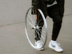 Cette photographie met en scène un **sujet principal flou**, partiellement visible : une personne en mouvement tenant une roue de vélo. La **composition** est simple, axée sur le bas du corps et la roue, suggérant un geste dynamique. Le **cadrage** est serré, centré sur les détails techniques de la roue et les pieds chaussés de baskets.La **lumière** semble naturelle, avec des contrastes modérés. Les **couleurs dominantes** sont neutres : gris (sol), noir (vêtements), et blanc/bleu (baskets). Le style est **documentaire et minimaliste**, évoquant une ambiance **urbaine et moderne**, presque abstraite par son flou.