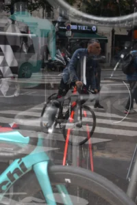 Cette photographie montre un cycliste comme sujet principal, reflété dans une surface vitrée. La composition joue sur la symétrie et le reflet, créant une impression de mouvement et de duplication.Le cadrage est serré sur le vélo et le cycliste, avec des éléments urbains en arrière-plan. La lumière est naturelle, légèrement tamisée, avec des ombres douces. Les couleurs dominantes sont les teintes froides (bleu turquoise du vélo, gris de la ville) et quelques touches chaudes (rouge du cadre).Le style est réaliste et urbain, évoquant une ambiance dynamique et moderne dans un cadre citadin.