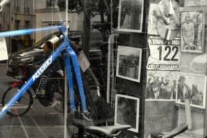 Cette photographie montre principalement un vélo bleu garé devant une vitrine. La composition inclut aussi des affiches anciennes en noir et blanc, dont une avec le numéro "122". Le cadrage est serré, mettant en valeur le vélo et les affiches en arrière-plan.La lumière semble naturelle, avec des contrastes doux. Les couleurs dominantes sont les tons bleus du vélo et les noirs et blancs des affiches. Le style est urbain et rétro, évoquant une ambiance mélancolique et nostalgique, mêlant modernité et passé.