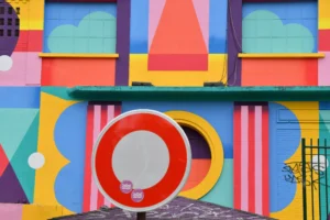 Cette photographie montre un mur aux formes géométriques colorées et stylisées. Le sujet principal est une composition abstraite et dynamique, mêlant cercles, triangles et rectangles. Le cadrage est serré, mettant en valeur les détails des formes.Les couleurs dominantes sont vives : bleu, jaune, rouge, orange et vert. La lumière semble naturelle, éclairant uniformément la surface sans ombre marquée. Le style est moderne et artistique, évoquant l’art abstrait ou le pop art. L’ambiance est joyeuse et énergique, avec une touche de fantaisie et de créativité.