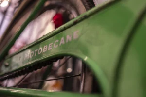 Cette photographie met en scène une partie d’un véhicule vintage, probablement une moto ou une voiture ancienne, de marque **S.A. MOTOBECAINE**.Le **sujet principal** est le flanc vert du véhicule, avec son nom partiellement visible. La **composition** est serrée, avec un **cadrage rapproché** qui isole la partie métallique et chromée. La **lumière** semble naturelle, mettant en valeur les détails et les reflets. Les **couleurs dominantes** sont le **vert profond** et le **noir**, avec des touches de chrome brillant.Le style est **documentaire et esthétique**, évoquant une ambiance **rétro et industrielle**, presque nostalgique.