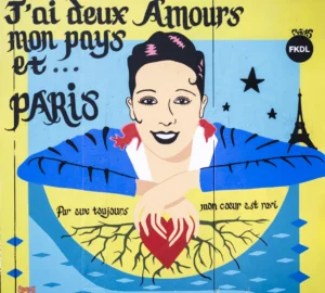 Cette photographie montre une affiche colorée mettant en scène une femme stylisée, coiffée et vêtue à la mode des années 1930-1940. Le sujet principal est une citation en français : *« J’ai deux Amours, mon pays et... Paris »*, avec des éléments comme la Tour Eiffel et un cœur rouge.La composition est dynamique, avec des couleurs vives (bleu, jaune, rouge) et un style rétro-art déco. Le cadrage est serré sur le visage et les épaules de la femme. La lumière semble artificielle, mettant en valeur les contours. L’ambiance est festive et nostalgique, mêlant patriotisme et amour pour Paris.