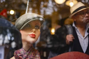 Cette photographie met en scène une tête de mannequin féminin comme sujet principal, vêtue d’un chapeau gris et d’une robe à motifs floraux. Le cadrage est serré, centré sur le mannequin, avec en arrière-plan des personnes floues portant des chapeaux.La lumière est douce et tamisée, créant une ambiance chaleureuse et légèrement vintage. Les couleurs dominantes sont les tons pastel (gris, rose pâle) et les motifs floraux, renforçant un style rétro ou années 1940-1950. L’ambiance évoque un marché aux puces ou une boutique de vêtements anciens.