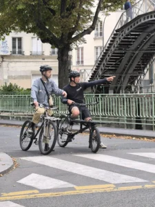 Cette photographie montre deux personnes à vélo, dont l’une semble guider l’autre sur un vélo tandem. Le sujet principal est ce duo en mouvement.La composition met en valeur la rue urbaine avec une perspective légèrement basse, encadrée par une grille et des escaliers en arrière-plan. Le cadrage est serré, capturant les détails des vélos et des vêtements des cyclistes.La lumière est naturelle et semble douce, typique d’une journée ensoleillée. Les couleurs dominantes sont les tons neutres (gris des vêtements, métal des vélos) et les verts des arbres, créant une ambiance à la fois dynamique et paisible, typique d’une balade urbaine.