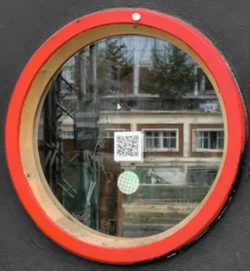 Cette photographie montre un miroir rond aux contours en bois et bordure rouge. Le sujet principal est la réflexion d’un environnement extérieur : un bâtiment, des arbres et un QR code collé sur la vitre.La composition est simple et centrée, avec un cadrage serré sur le miroir. La lumière semble naturelle, bien que légèrement atténuée, créant une ambiance douce et légèrement floue. Les couleurs dominantes sont les tons neutres (beige, gris) du bâtiment et les touches de vert des arbres, contrastées par le rouge vif du cadre. Le style est épuré et réaliste, évoquant une ambiance calme et introspective.