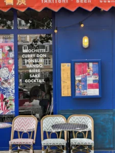Cette photographie montre l'extérieur d'un bar ou restaurant nommé "Dango". Le sujet principal est l'affiche lumineuse bleue listant des plats et boissons, comme "Curry Don" et "Cocktail".La composition inclut des chaises en métal et plastique disposées devant, ainsi que des affiches colorées sur les murs. Le cadrage est serré, mettant en valeur les détails du menu et de la façade.Les couleurs dominantes sont le bleu vif et des touches de rouge et blanc. La lumière semble naturelle, bien que renforcée par l'éclairage artificiel de l'affiche. L'ambiance est conviviale et moderne, évoquant un lieu branché et accueillant.