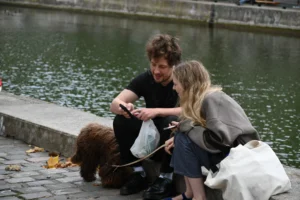 Cette photographie montre deux personnes assises près d’un plan d’eau, semblant interagir avec un chien. Le sujet principal est cette scène de complicité entre les humains et l’animal.La composition est naturelle, avec un cadrage serré sur les protagonistes et un arrière-plan flou centré sur l’eau. La lumière semble douce et naturelle, probablement en journée. Les couleurs dominantes sont des tons neutres (gris, bleu de l’eau) et quelques touches de couleurs vives comme le jaune des feuilles mortes.Le style est réaliste et spontané, créant une ambiance paisible et chaleureuse.