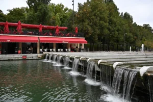 Cette photographie met en scène une série de **chutes d’eau en cascade** comme sujet principal. La composition repose sur une **perspective linéaire**, avec des bassins alignés et un cadrage serré sur les cascades.La lumière est **naturelle et douce**, probablement en journée, créant des reflets sur l’eau. Les **couleurs dominantes** sont les teintes **vertes** des arbres, le **rouge vif** du bâtiment en arrière-plan et le **bleu grisâtre** de l’eau.Le style est **réaliste et photographique**, avec une ambiance **calme et apaisante**, évoquant un espace de détente en milieu urbain ou naturel.