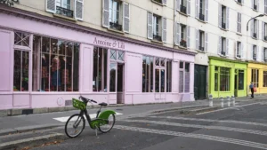 Cette photographie met en scène un vélo vert garé devant des bâtiments colorés. Le sujet principal est ce vélo, positionné sur un trottoir.La composition est simple et équilibrée, avec une perspective légèrement en contre-plongée. Le cadrage est serré, centré sur le vélo et les façades.Les couleurs dominantes sont pastel : rose, vert et jaune, apportant une ambiance chaleureuse et apaisante. La lumière semble naturelle, douce, typique d’une journée ensoleillée.Le style est urbain et contemporain, évoquant une ambiance paisible et esthétique dans une ville européenne.