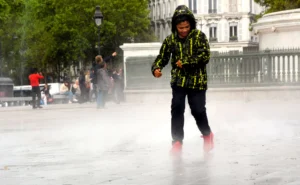 Cette photographie capture un enfant comme sujet principal, vêtu d’un manteau coloré et de bottes, courant sous une pluie battante. La composition montre un cadrage dynamique, centré sur l’enfant, avec des éléments urbains en arrière-plan (bâtiments, arbres, passants).La lumière est tamisée par la pluie, créant une ambiance floue et humide. Les couleurs dominantes sont les tons gris de l’eau et du ciel, contrastés par les teintes vives du manteau jaune et vert de l’enfant. Le style est documentaire et spontané, évoquant une scène de vie quotidienne. L’ambiance est à la fois joyeuse et mélancolique, reflétant la liberté malgré la pluie.