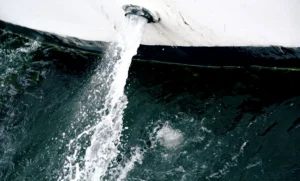 Cette photographie capture un **geyser naturel** ou une chute d’eau souterraine jaillissant dans une mer ou un lac.**Sujet principal** : Le jet d’eau central, dynamique et puissant.
**Composition** : Cadrage serré, centré sur le flux ascendant, avec une symétrie verticale.
**Lumière** : Contrastée, avec des ombres marquées et une lumière naturelle, probablement en journée.
**Couleurs dominantes** : Noir et blanc, avec des nuances de gris, accentuant les textures et contrastes.
**Style** : Documentaire et artistique, évoquant une scène naturelle brute.
**Ambiance** : Mystérieuse et puissante, soulignant la force de la nature.